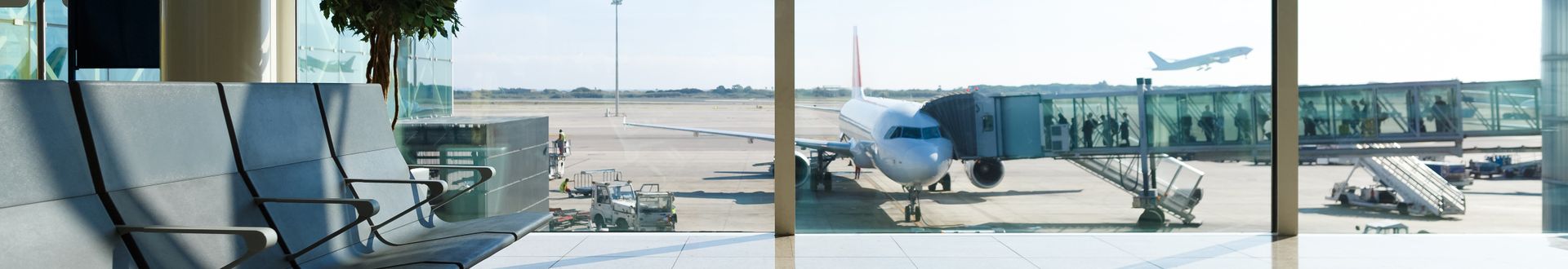 Moderne Wartezone im Flughafenterminal mit Blick auf das Vorfeld, ein angedocktes Passagierflugzeug an der Fluggastbrücke sowie ein startendes Flugzeug im Hintergrund