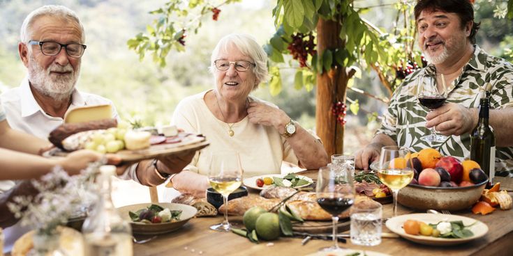 Mehrere Personen sitzen an einem gedeckten Holztisch im Freien unter Weinreben, mit Tellern, Gläsern und einer Obstschale mit Pfirsichen und Trauben.