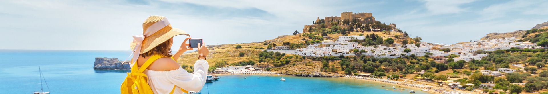 Frau mit Sonnenhut und gelbem Kleid fotografiert Küstenstadt mit Burg auf Hügel und blauem Meer im Hintergrund.