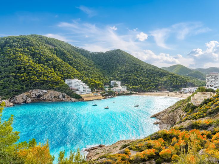 Bucht von Cala Llonga auf Ibiza mit türkisblauem Wasser, umgeben von bewaldeten Hügeln und mehreren weißen Gebäuden am Ufer.