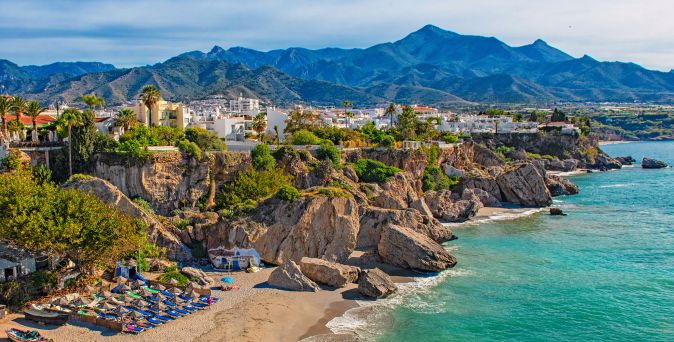 Küstenansicht mit Felsen, Sandstrand und Liegen. Im Hintergrund sind Berge und Gebäude sichtbar.