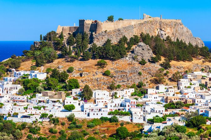 Blick auf die Akropolis von Rhodos auf einem bewaldeten Hügel über einem Dorf mit weißen Häusern und blauem Himmel.