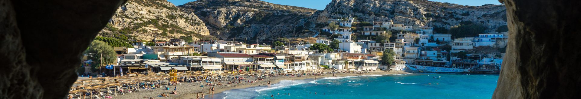 Blick aus einer Höhle auf den Strand von Matala auf Kreta mit mehreren Sonnenschirmen, Menschen und weißen Gebäuden am Hang.
