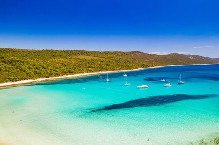 Blick über den Sakarun Beach auf Dugi otok in Kroatien mit flachem türkisfarbenem Wasser, vor Anker liegenden Segelbooten und der naturbelassenen Küstenlandschaft der Adria