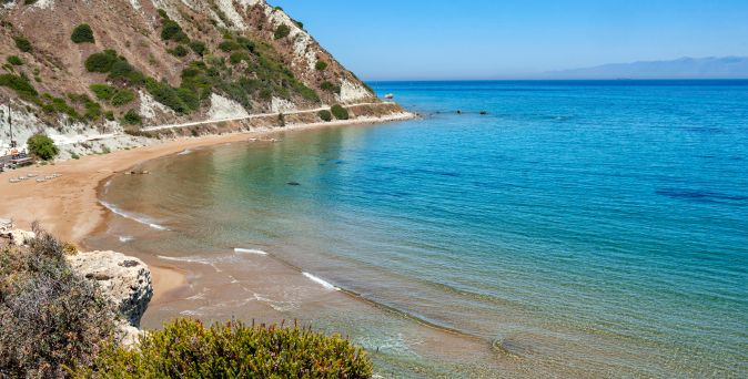 Kleiner Sandstrand mit klarem, ruhigem Wasser, umgeben von felsigen Klippen und vereinzelter Vegetation.