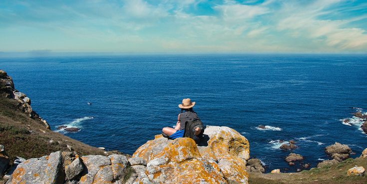 Person mit Hut und Rucksack sitzt auf Felsen und blickt auf das weite Meer mit vereinzelten Felsen im Wasser.