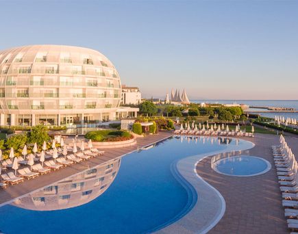 Großes, rundliches Hotelgebäude neben einem geschwungenen Außenpool mit Reihen von Liegestühlen und Sonnenschirmen am Ufer