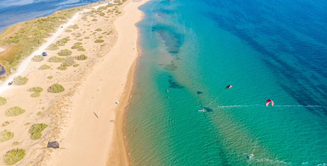 Luftaufnahme des Issos-Strands auf Korfu mit Sanddünen links und klarem, türkisfarbenem Wasser rechts, in dem mehrere Kitesurfer aktiv sind.