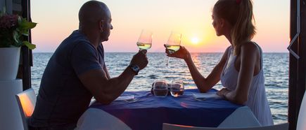 Ein Paar sitzt in einem Restaurant direkt am Meer bei Sonnenuntergang und halten Weingläser