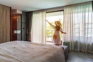 Frau mit Hut und weißem Kleid steht in Hotelzimmer vor großem Fenster und blickt nach draußen