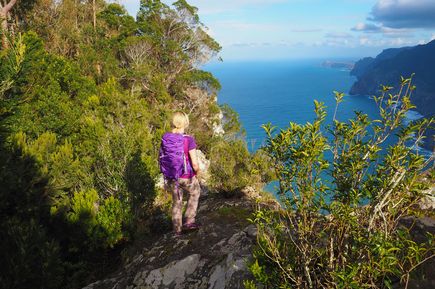 Person mit Rucksack steht auf dem Wanderweg Vereda do Larano auf Madeira und blickt über die bewaldete Steilküste und den Atlantik