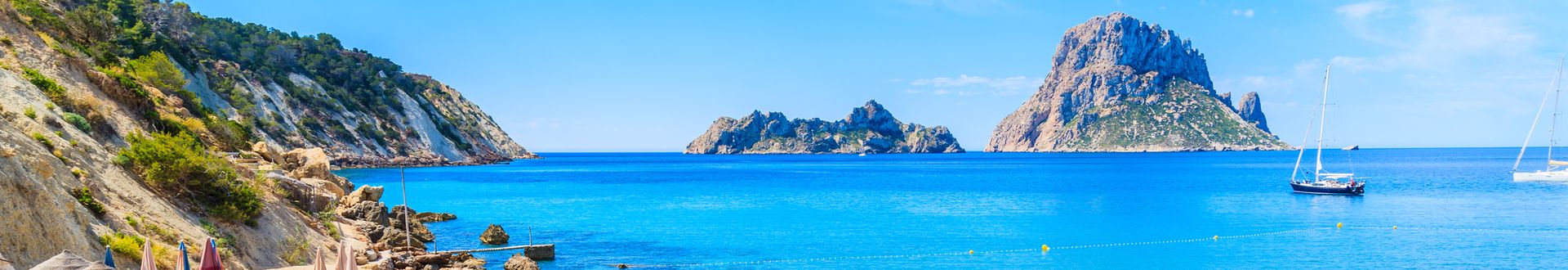 Blick auf die Bucht Cala d’Hort auf Ibiza mit klarem, blauem Wasser, Felsen, grünen Klippen und Segelbooten vor der Insel Es Vedrà
