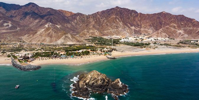 Blick auf einen Strand mit Felsen im Wasser und dahinter eine Siedlung vor bergiger Wüstenlandschaft in Fujairah