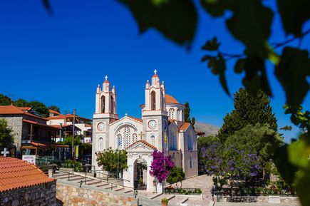 Kirche mit zwei Türmen und Kuppel, umgeben von Bäumen und Gebäuden, unter klarem blauem Himmel.