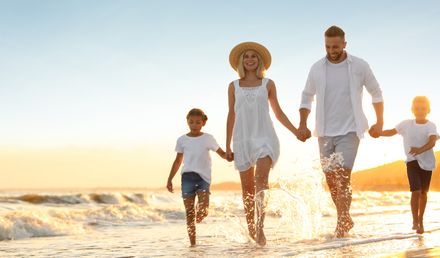 Familie mit zwei Kindern läuft am Strand bei Sonnenuntergang, alle tragen helle Kleidung