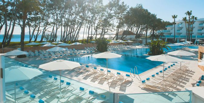 Großer Außenpool des Iberostar Waves Playa de Muro auf Mallorca mit zahlreichen Liegen und weißen Sonnenschirmen, umgeben von hohen Bäumen und mit Blick auf das Meer sowie die modernen Hotelgebäude