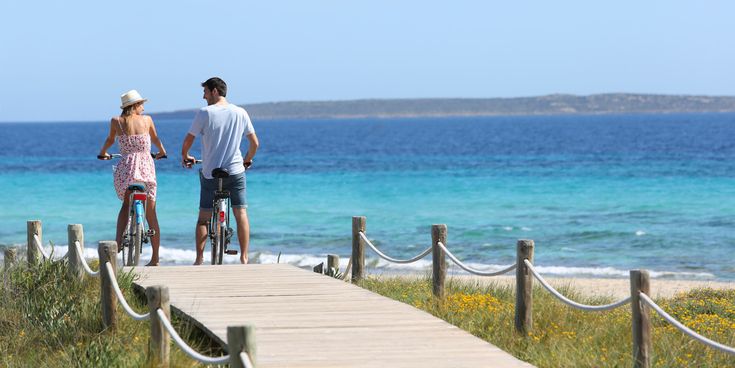 Ein Paar fährt mit Fahrrädern über einen Holzsteg am Strand und blickt auf das türkisfarbene Meer