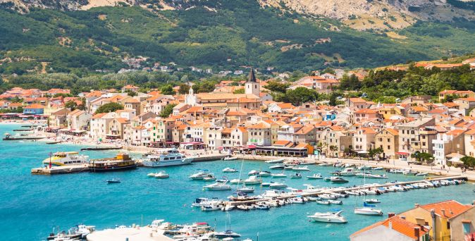 Blick auf den Küstenort Baška auf der Insel Krk in Kroatien mit Hafen, zahlreichen Booten, Uferpromenade und Bergen im Hintergrund