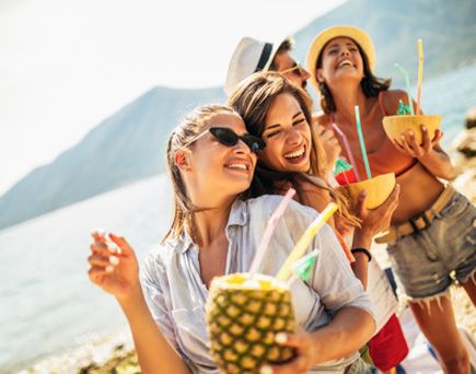 Drei Frauen am Strand, eine hält eine Ananas mit Strohhalm, eine andere trägt einen Strohhut, alle tragen sommerliche Kleidung.