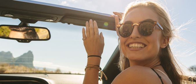 eine Person sitzt in einem Auto mit erhobenen Händen, im Hintergrund Berge und blauer Himmel.