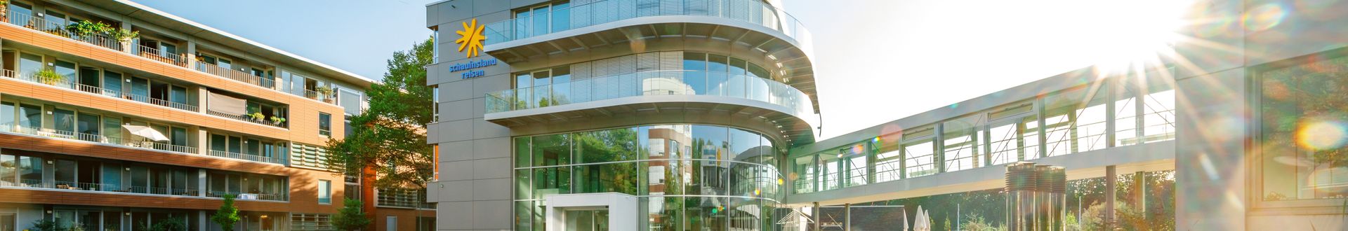 Außenansicht von Gebäude 3 des Hauptsitzes von schauinsland-reisen mit moderner Glasfassade, abgerundeten Balkonen, Firmenlogo, begrüntem Innenhof und verbindendem Glasgang zwischen den Gebäuden bei Sonnenschein