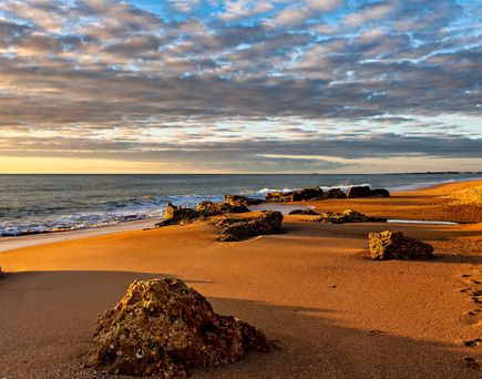 Strand mit Felsen und goldfarbenem Sand bei Sonnenuntergang unter bewölktem Himmel