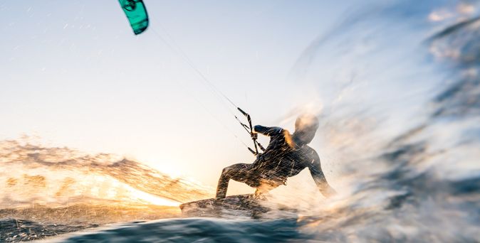 Ein Windsurfer in einem schwarzen Neoprenanzug reitet auf einer Welle, während ein Kiteschirm in Blau und Schwarz über ihm schwebt