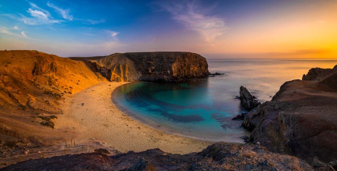 Panoramablick auf eine Küstenlandschaft mit einer geschwungenen Bucht, feinem Sandstrand und klarem, türkisfarbenem Wasser, umgeben von felsigen Ufern und sanften Hügeln.