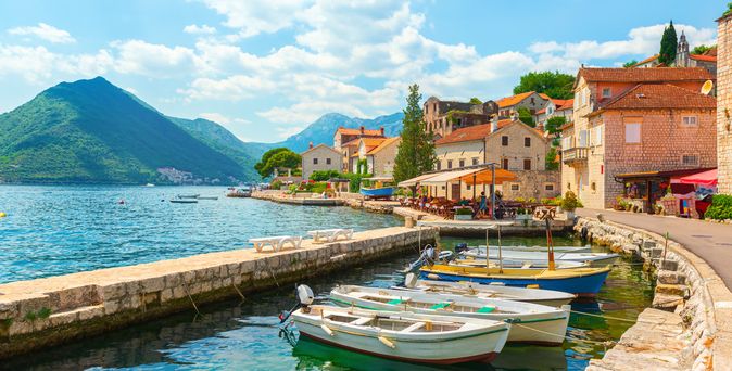 Kleiner Hafen mit mehreren kleinen Booten am Ufer eines klaren Gewässers, umgeben von Häusern mit roten Dächern und Bergen im Hintergrund