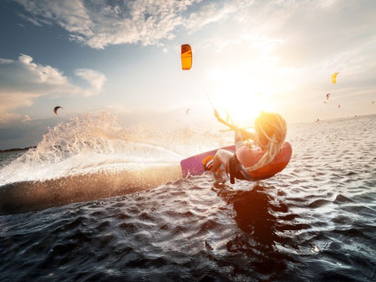 Kitesurfer gleitet dynamisch über das Wasser, während im Hintergrund mehrere Kites in der Luft schweben, Sonnenuntergangsstimmung