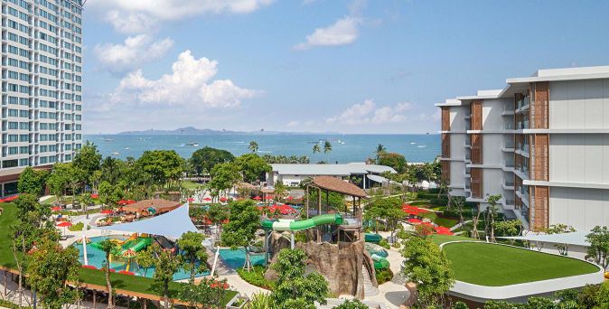 Blick auf ein Hotelresort mit Pool, Wasserrutschen, Spielplatz, Grünflächen und Meer im Hintergrund