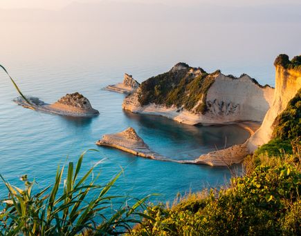 Blick auf die felsige Küste und das blaue Meer der Insel Korfu in Griechenland, mit grüner Vegetation im Vordergrund