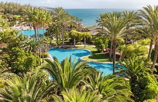 Großer Pool mit geschwungener Form, umgeben von Palmen und tropischer Vegetation, im Hintergrund Meer und bewölkter Himmel.