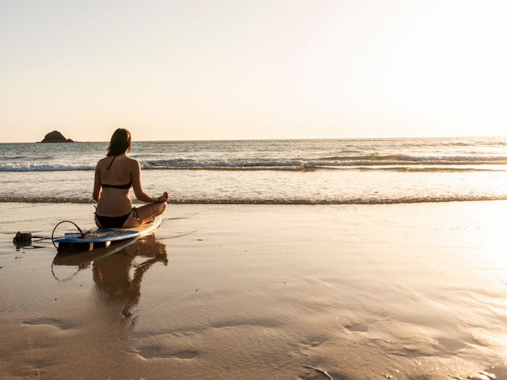 Frau im Bikini sitzt im Schneidersitz auf ihrem Surfbrett am Strand und blickt beim Sonnenuntergang aufs Meer