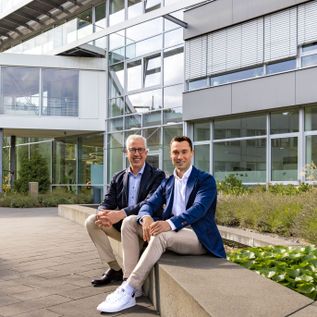 erald Kassner und Steffen Kassner sitzen gemeinsam im Außenbereich vor einem modernen Bürogebäude, beide in Businesskleidung, mit Glasfassade und Begrünung im Hintergrund