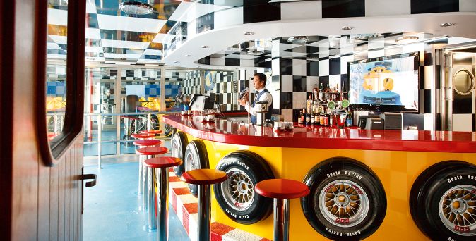 Die Bar Scuderia an Bord der Costa Pacifica mit rot gestalteter Rennsport-Theke, Felgen-Dekoration, Barhockern und großem Getränkeangebot in einem schwarz-weiß karierten Interieur
