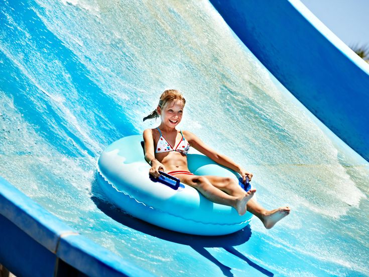 Mädchen in Bikini sitzt fröhlich in einem blauen Schwimmreifen auf einer Wasserrutsche