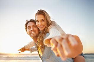Zwei Personen genießen am Strand den Sonnenuntergang, während der Mann die Frau auf dem Rücken trägt und beide die Arme fröhlich ausstrecken