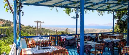 Überdachte Außenterrasse eines Restaurants mit blau-weißer Pergola, rot-weiß karierten Tischdecken und Holztischen mit Stühlen, im Hintergrund Blick auf grüne Hügel und das Meer.