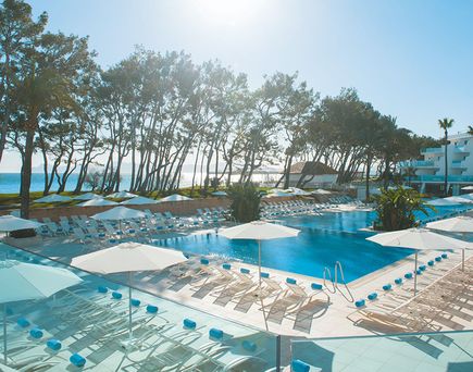 Großer Außenpool des Iberostar Waves Playa de Muro auf Mallorca mit zahlreichen Liegen und weißen Sonnenschirmen, umgeben von hohen Bäumen und mit Blick auf das Meer sowie die modernen Hotelgebäude