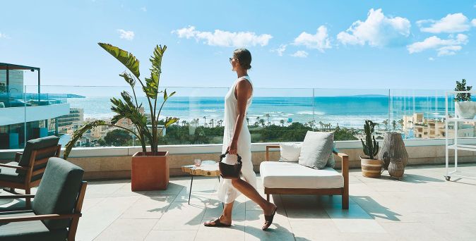Moderne Rooftop‑Terrasse des Iberostar Selection Llaut Palma mit Lounge-Möbeln und Blick auf das Mittelmeer, während eine Frau in Sommerkleidung vorbeigeht