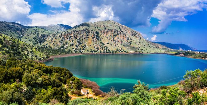Blick auf einen ruhigen See vor bewaldeten Hügeln und Bergen unter blauem Himmel auf Kreta, Griechenland