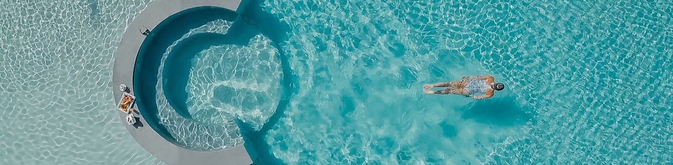 Eine Person schwimmt in einem klaren, blauen Pool. Am Rand des Pools liegen Handtücher und ein Getränk.