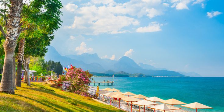 Strand von Kemer an der türkischen Riviera mit Sonnenschirmen, klarem türkisblauem Meer, grünen Palmen und Blick auf das Taurusgebirge