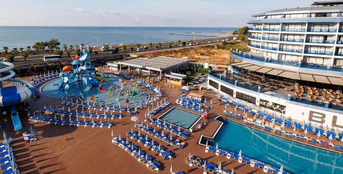 Blick auf mehrere Schwimmbecken mit Liegen und Sonnenschirmen neben einem Hotelgebäude und Palmen am Meer