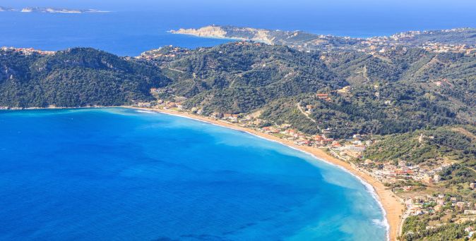Luftaufnahme einer geschwungenen Bucht mit Sandstrand, bewaldeten Hügeln und vereinzelten Gebäuden am Ufer.