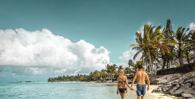 Paar in Badebekleidung hält Händchen und geht am Strand entlang, Palmen und ruhiges Meer im Hintergrund