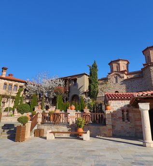 Steinerner Innenhof mit Bänken, Töpfen und blühendem Baum vor mehreren Gebäuden mit roten Ziegeldächern und Rundtürmen unter blauem Himmel.
