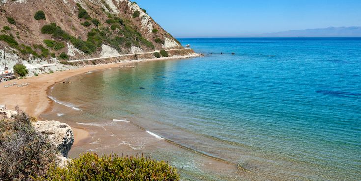 Kleiner Sandstrand mit klarem, ruhigem Wasser, umgeben von felsigen Klippen und vereinzelter Vegetation.