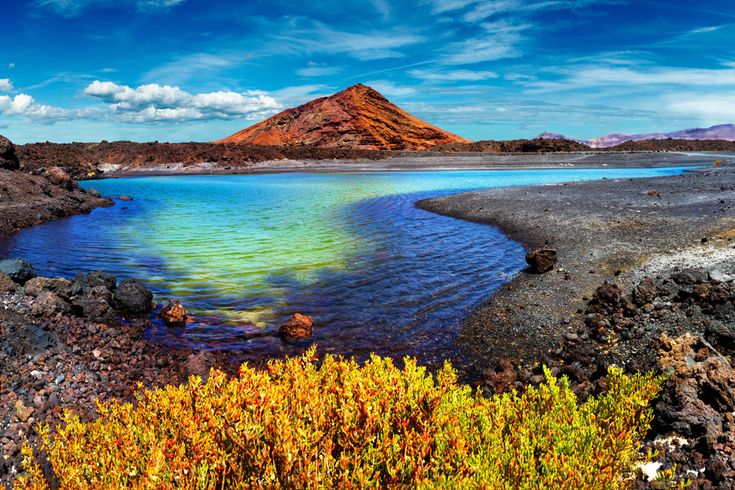 Vulkanlandschaft mit schwarzem Lavagestein, einem türkisfarbenen See und einem kegelförmigen Berg im Hintergrund unter blauem Himmel mit Wolken